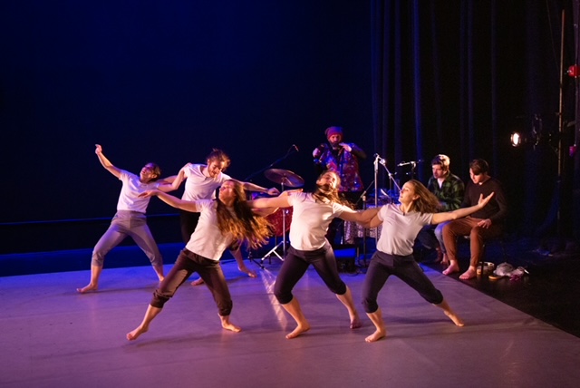 a group of five dancers wearing white t shirts and leggings dance together arm in arm in front of a band of musicians